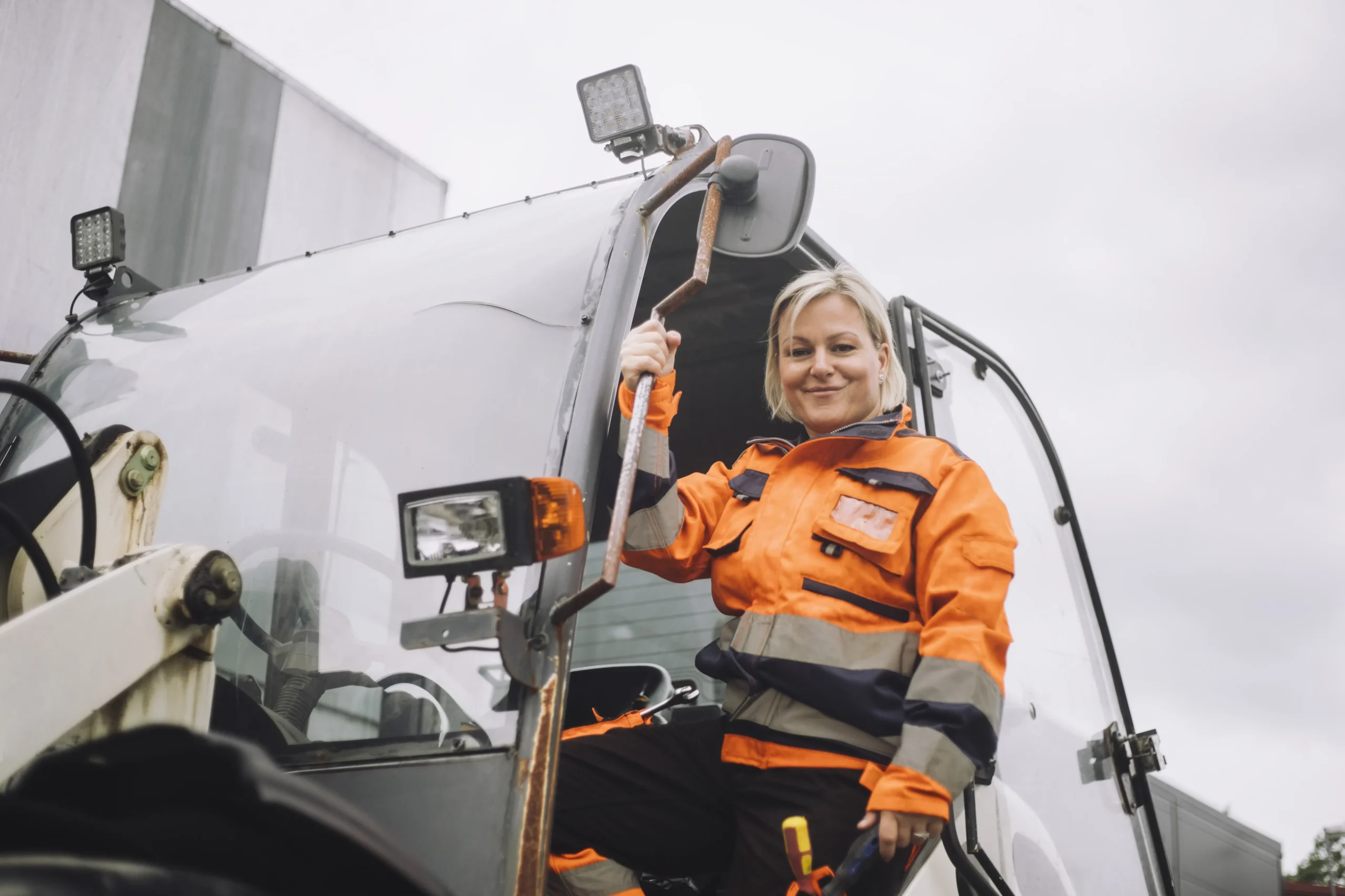 Vrouw in de deuropening van een vrachtwagen met een oranje overall aan.
