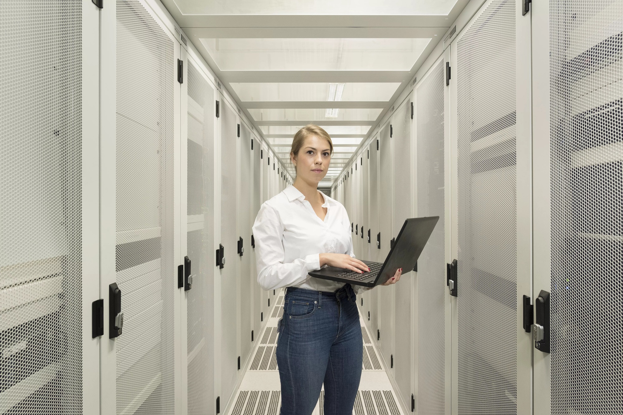 Een vrouw met een spijkerbroek en een witte blouse staat in een ruimte met diverse servers. Ze kijkt richting de camera en houdt een laptop vast.