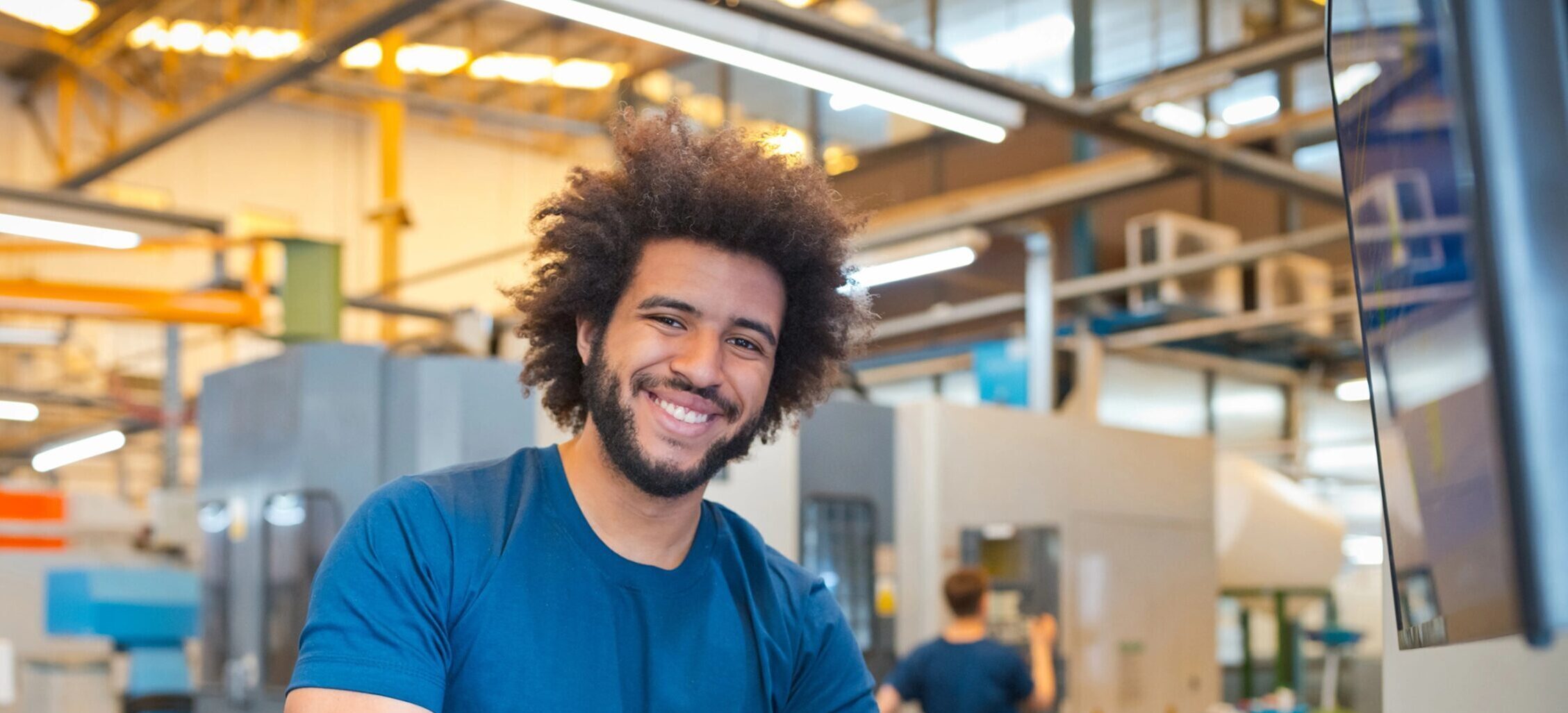 Een glimlachende man die in een fabriek achter een computer staat. Op de achtergrond staat een grote snijmachine.