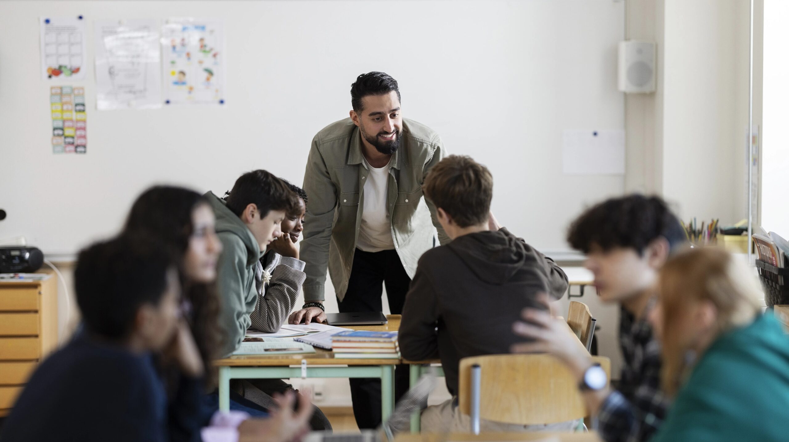 Een mannelijke leraar staat glimlachend aan tafel bij een groepje scholieren.