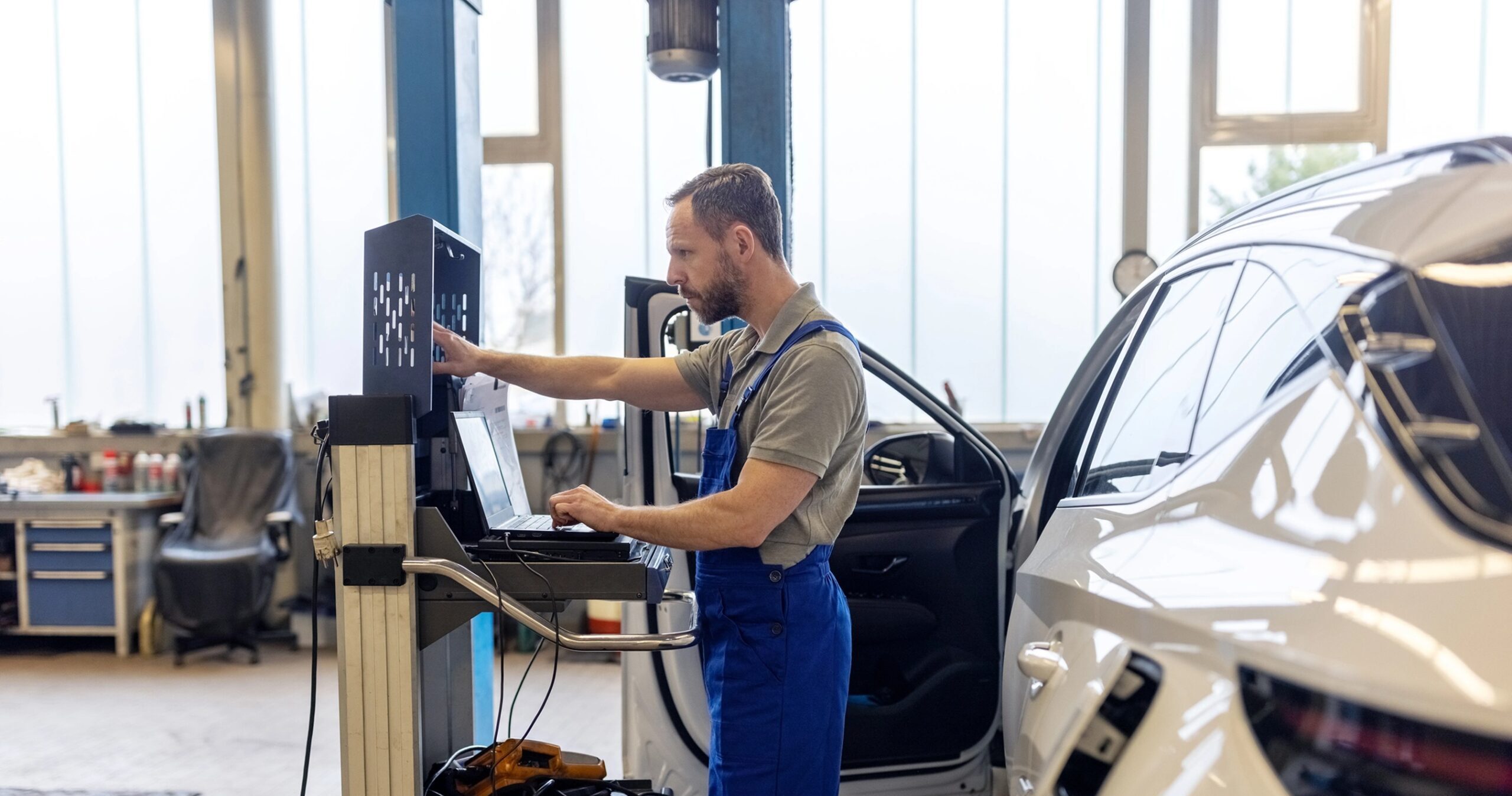 Een mannelijke monteur die met zijn rug richting een witte auto staat. Voor zich heeft hij een laptop staan om gegevens te verwerken.