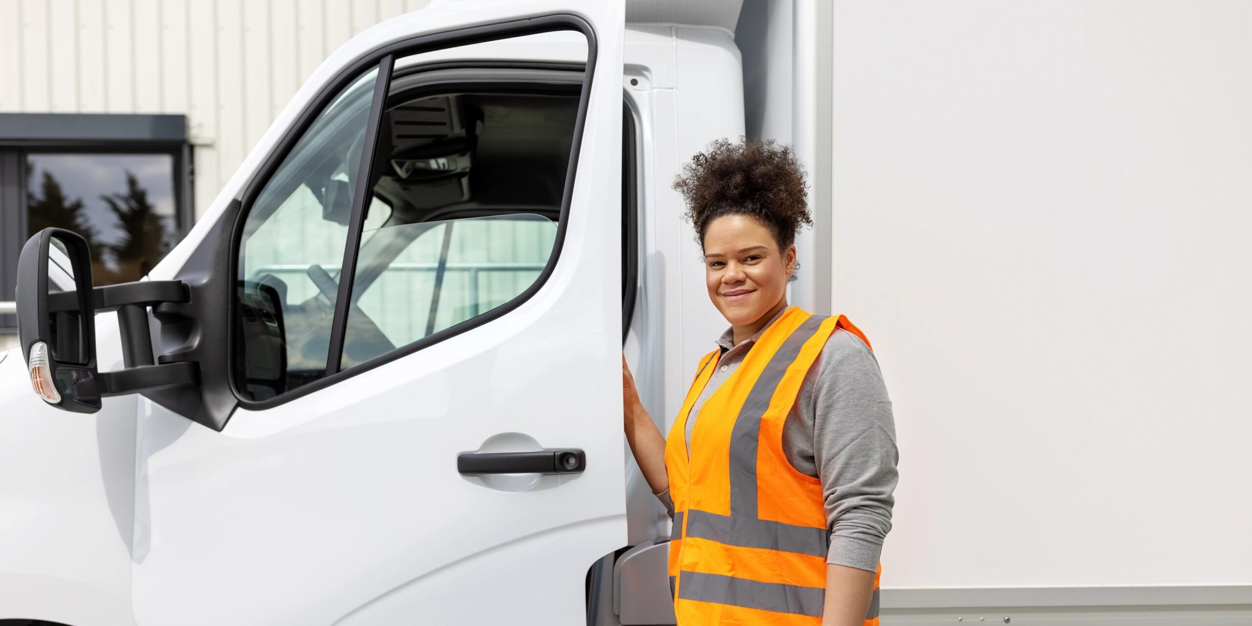 Een vrouw met een oranje hesje aan die de deur openhoud van een witte bakwagen.